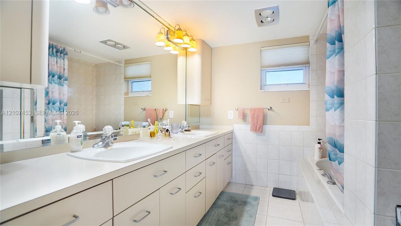 Bathroom featuring a full-width vanity with dual integrated sinks, extensive cabinetry, a large wall-mounted mirror, and tile-surround tub