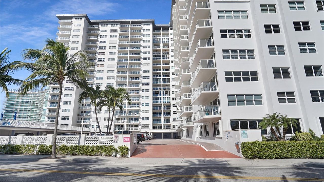 Multi-story high-rise exterior featuring a reinforced concrete facade, private balconies with glass railings, and ground-level paved parking