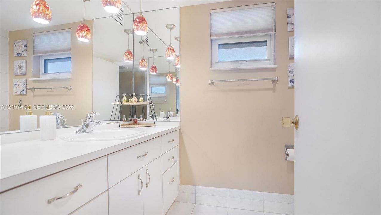 White vanity with integrated countertop and sink, large wall-to-wall mirror, multi-pendant lighting, tile flooring, and a window with a built-in shade