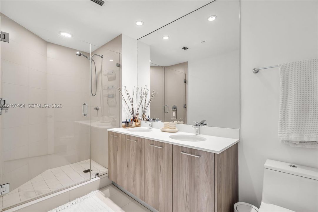 Double vanity featuring wood-finish cabinetry, solid surface countertops, dual undermount sinks, and a frameless glass-enclosed shower with tiled walls