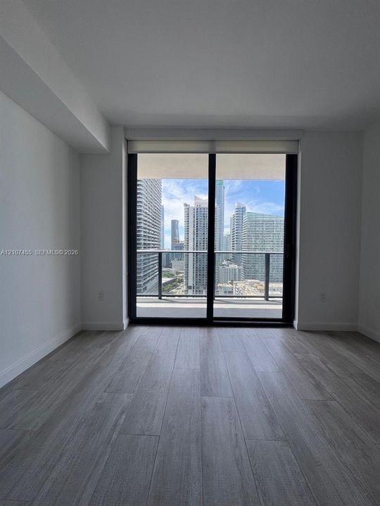 Floor-to-ceiling sliding glass door, wood-finish flooring, private balcony with metal railing, and urban skyline backdrop
