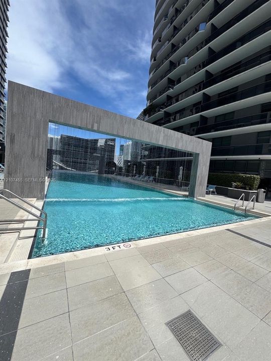 Outdoor swimming pool featuring a stone-clad waterfall feature, tiered edges, metal handrails, and paved deck surfacing