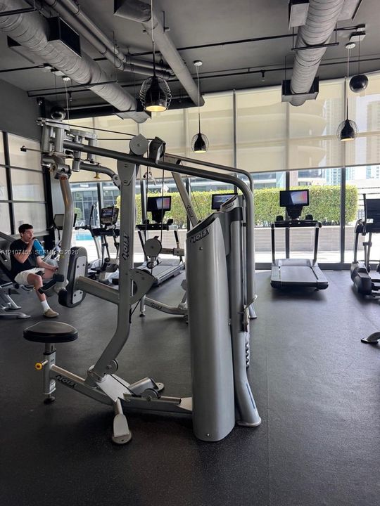 Fitness facility featuring floor-to-ceiling windows, high-contrast industrial-style ceiling with exposed ductwork, rubberized flooring, and pendant lighting