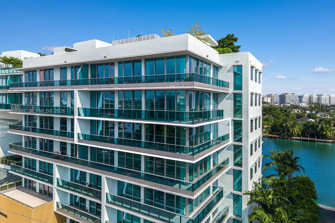 Modern multi-story building featuring white stucco facade, expansive floor-to-ceiling windows, wrap-around glass-railed balconies, and a rooftop terrace