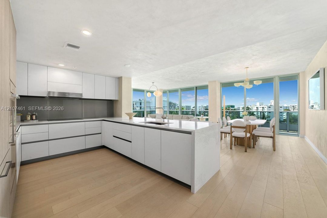 Contemporary kitchen featuring white handleless cabinetry, a substantial quartz-topped center island with integrated sink, stainless steel range hood, and light-toned wood-finish flooring, complemented by floor-to-ceiling glass wall panels