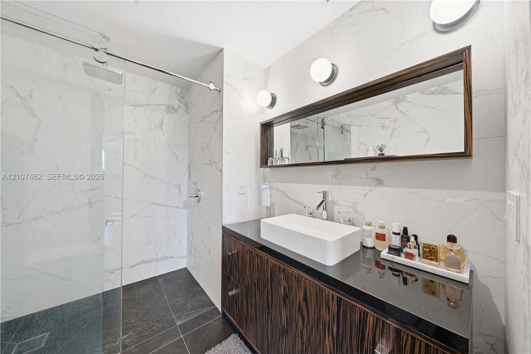 Bathroom featuring a wall-mounted vanity with a dark wood-finish cabinet and dark stone countertop, rectangular vessel sink, floor-to-ceiling large-format tile surround, frameless glass shower enclosure, and integrated mirror cabinetry