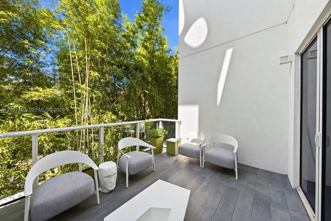 Private balcony featuring gray wood-finish decking, a white metal railing with glass panels, and mature bamboo landscaping