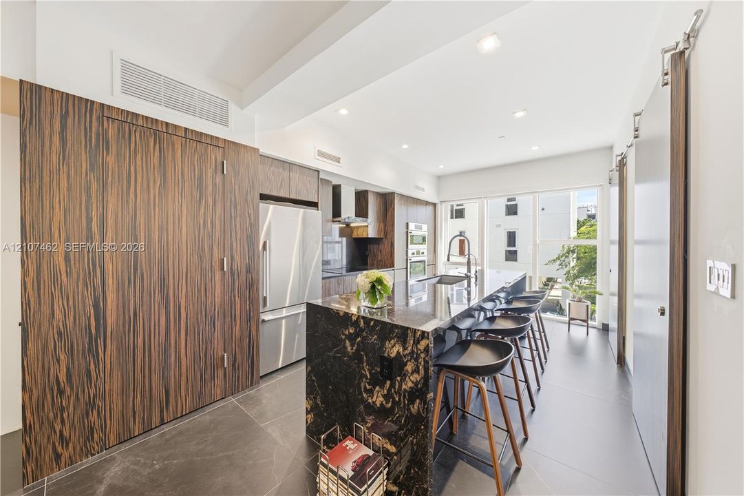 Kitchen featuring full-height wood-finish cabinetry, a waterfall-edge stone island, built-in stainless steel appliances, large-format tile flooring, and recessed ceiling lighting
