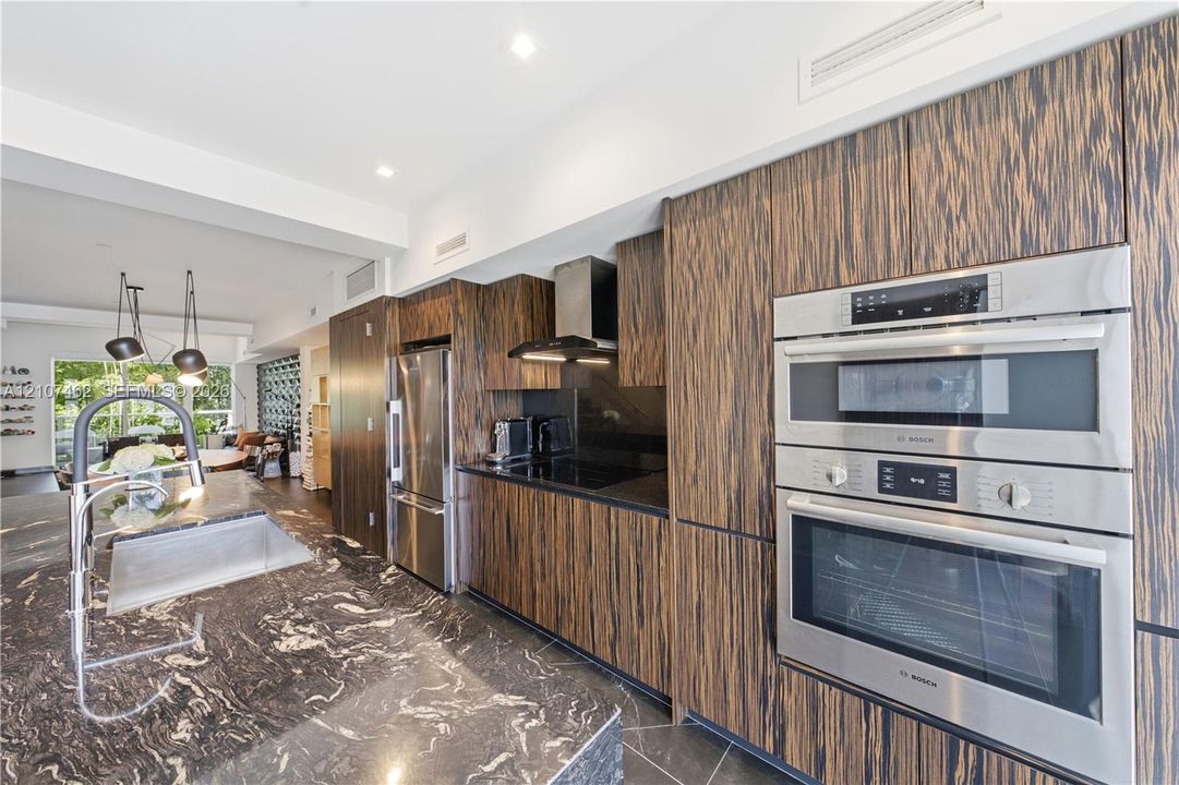 Kitchen featuring horizontal-grain wood cabinetry, integrated stainless steel appliances, dark stone countertops, stone flooring, and recessed ceiling lighting