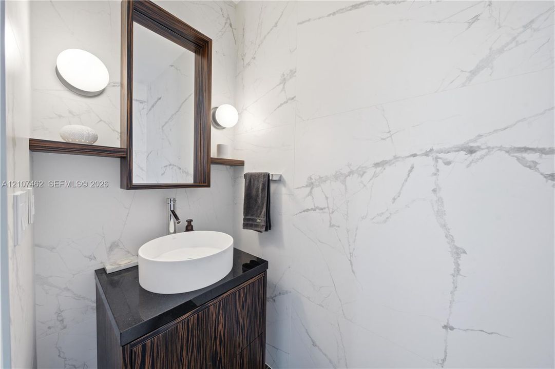 Dark wood-finish vanity with a stone countertop, white oval vessel sink, wall-mounted mirror with integrated shelving, and floor-to-ceiling stone-style wall tile