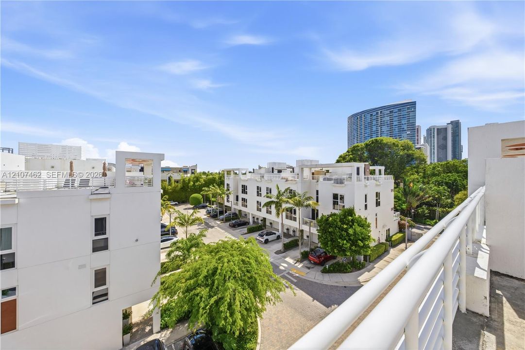 Elevated outdoor balcony with white railings overlooking a residential complex featuring multi-level structures, paved driveways, and mature landscaping