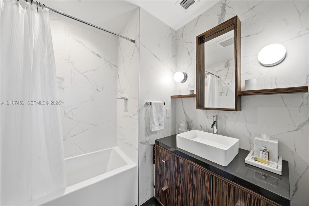 Bathroom featuring large-format wall tiling, a dark stone vanity countertop, a rectangular vessel sink, a wood-finish vanity cabinet, and wall-mounted vanity lighting