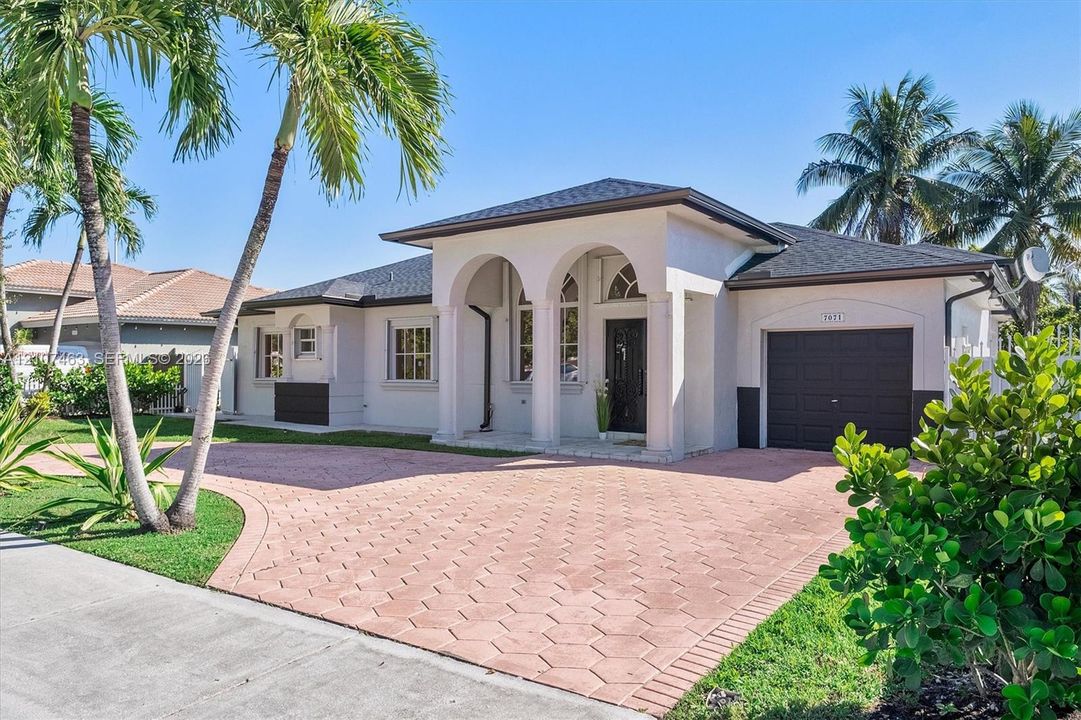 Single-story exterior featuring a covered arched entryway, hexagonal paved driveway, dark-toned garage door, and mature palm trees