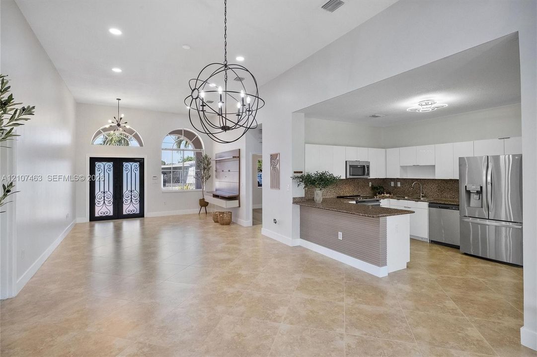 Open-concept interior featuring large-format tile flooring, arched windows with decorative double entry doors, a spherical multi-light chandelier, and a kitchen equipped with flat-panel cabinetry and stainless steel appliances