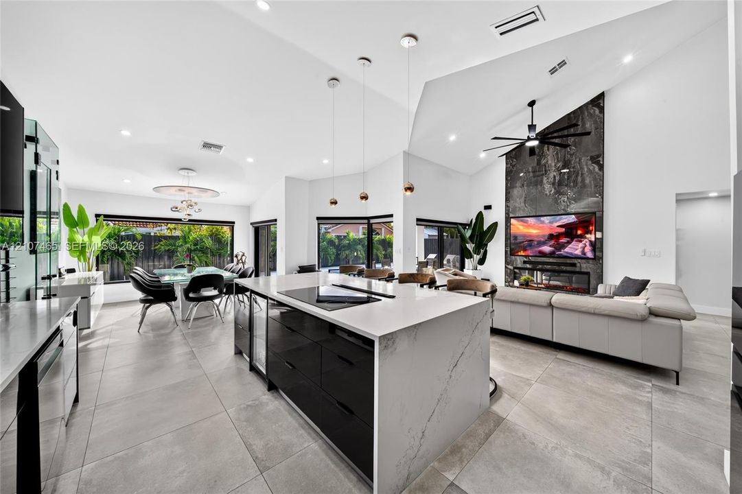 Open-concept floor plan featuring high vaulted ceilings, large-format tile flooring, a center island with stone-finish waterfall countertop, floor-to-ceiling stone-finish fireplace surround, and modern multi-light pendant fixtures
