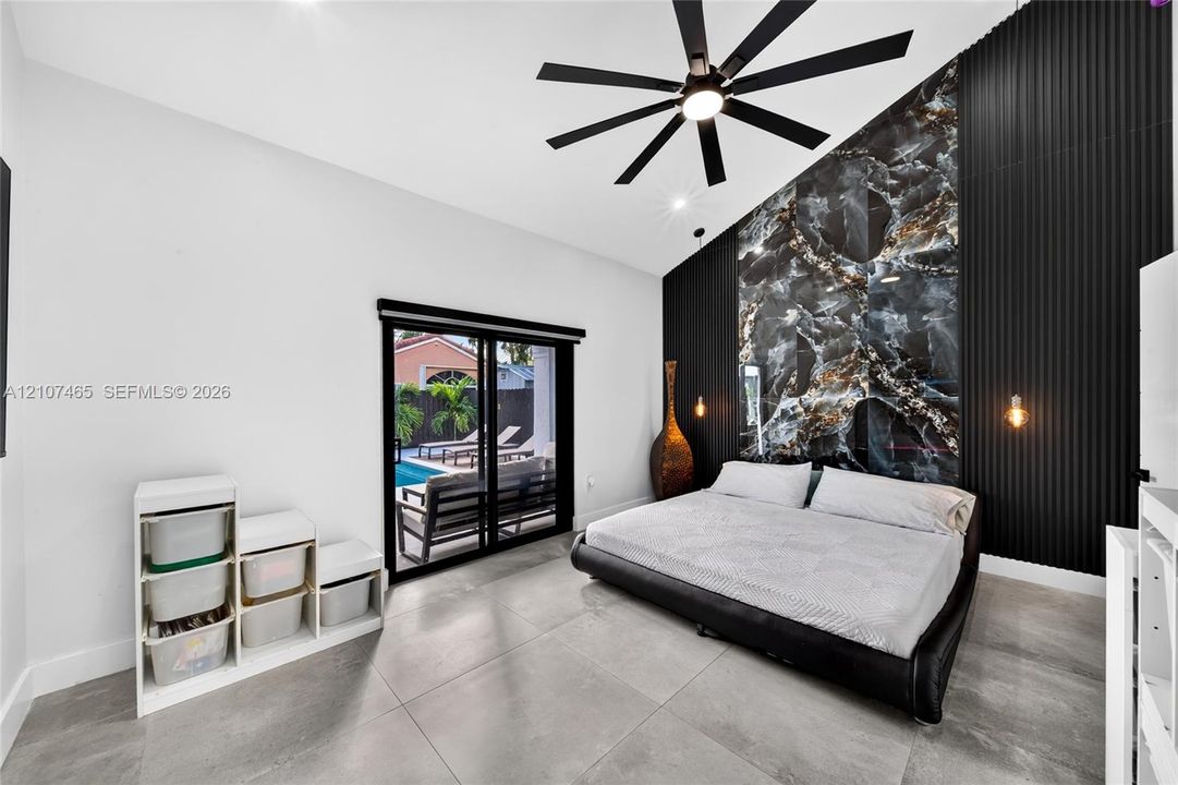 Vaulted ceiling room featuring large-format tile flooring, a statement accent wall with dark stone-finish cladding and vertical slats, a black multi-blade ceiling fan, and sliding glass doors