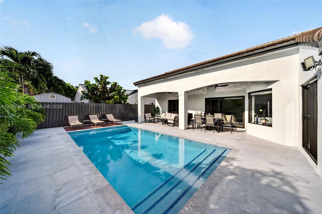 Rear exterior featuring a covered patio, rectangular swimming pool with built-in stairs, neutral-toned stone paver decking, and privacy fencing