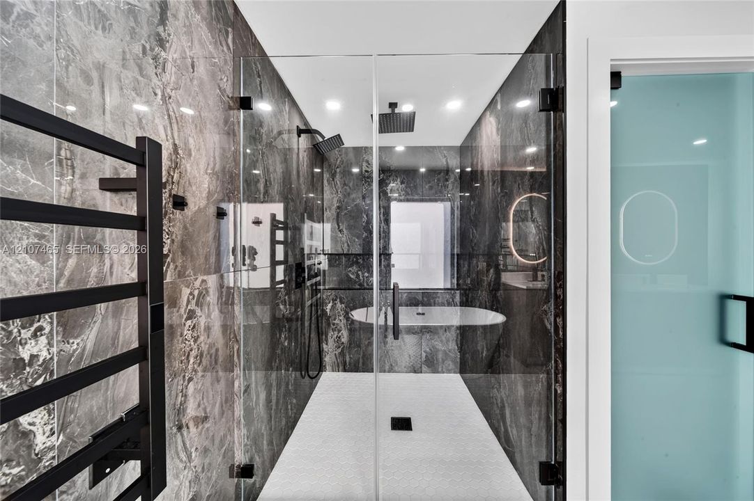 Walk-in shower featuring floor-to-ceiling stone-patterned wall tile, dual rainfall showerheads, a glass enclosure, a freestanding soaking tub, and a matte black wall-mounted towel warmer