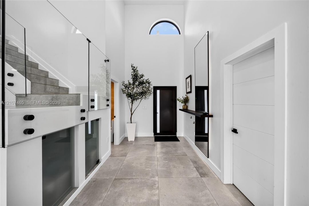 Foyer featuring double-height ceilings, large-format tile flooring, a glass-panel staircase railing, a semicircular clerestory window, and a modern entrance door with a vertical glass insert