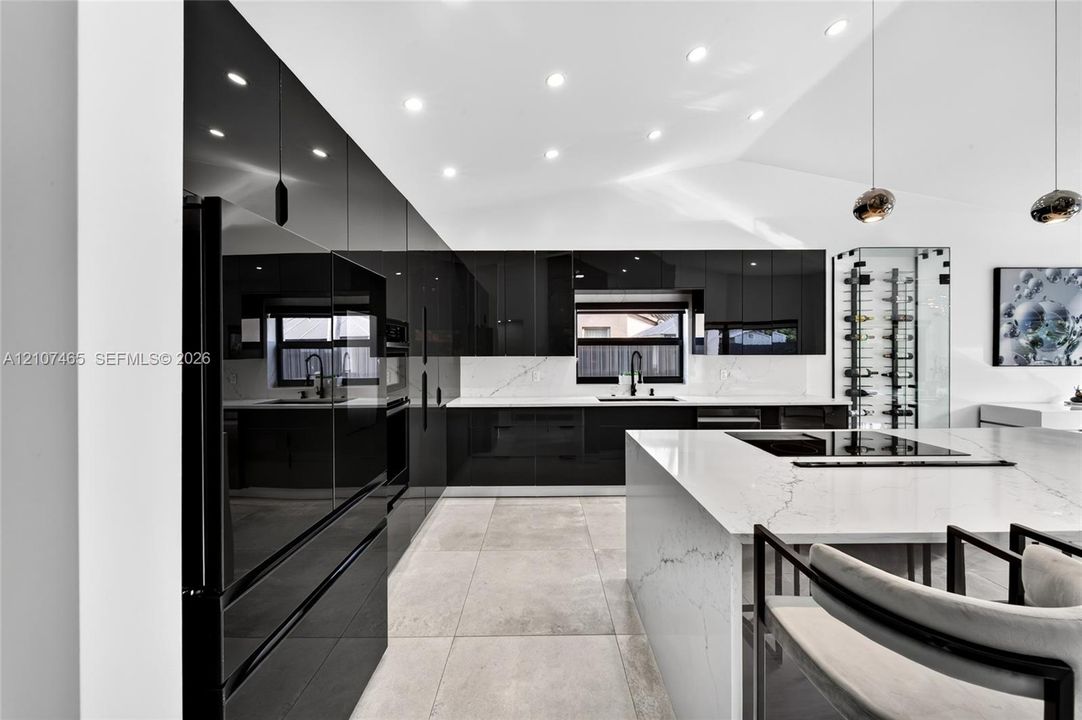 Modern kitchen featuring high-gloss black cabinetry, white stone countertops with veining, a central island with an integrated cooktop, large-format tile flooring, and a built-in glass-encased wine storage display