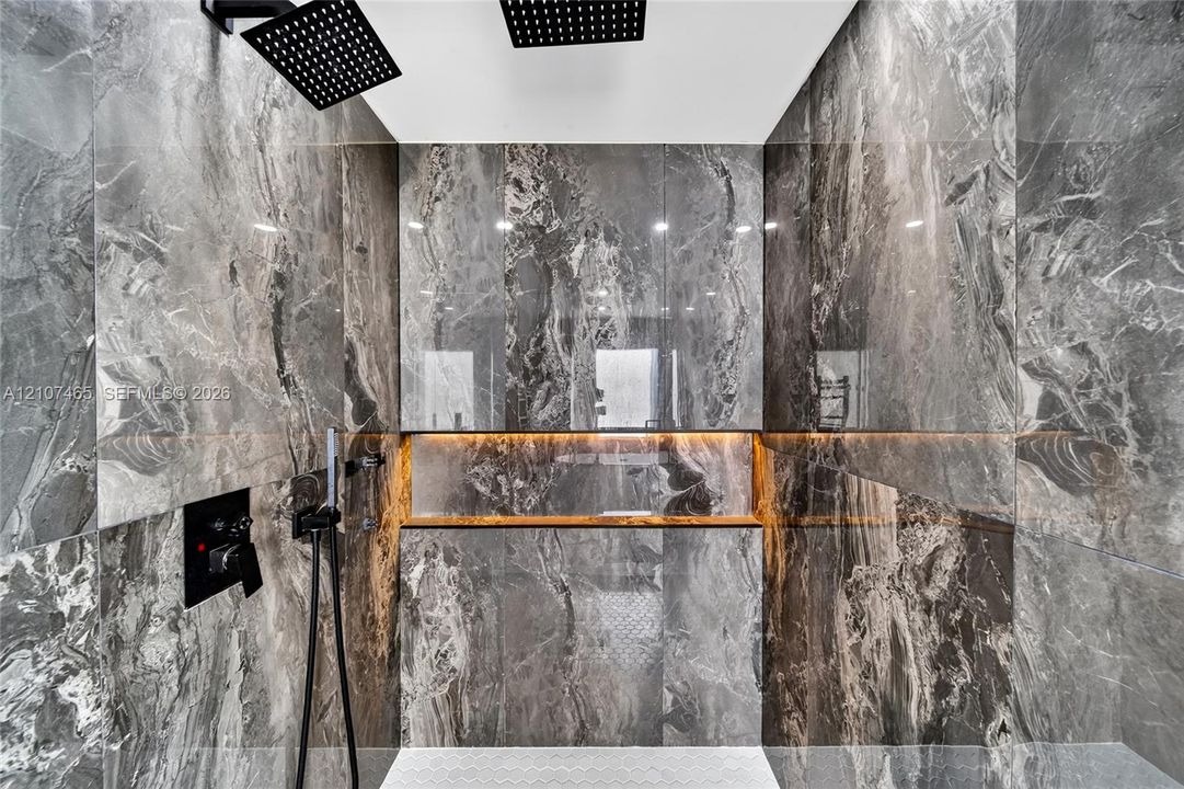 Walk-in shower featuring large-format stone-finish wall tile, dual overhead rainfall showerheads, handheld shower wand, integrated LED-lit recessed shelving, and matte black plumbing fixtures