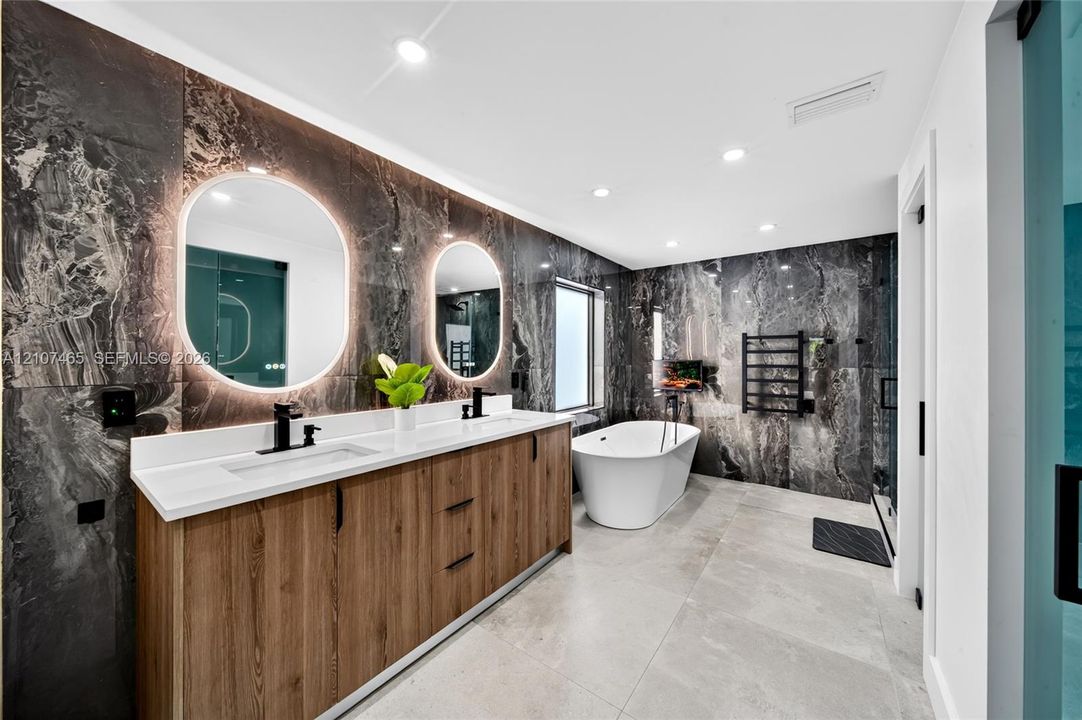 Dual-sink vanity with wood-finish cabinetry, stone-tiled accent walls, freestanding soaking tub, backlit oval mirrors, and matte black fixtures