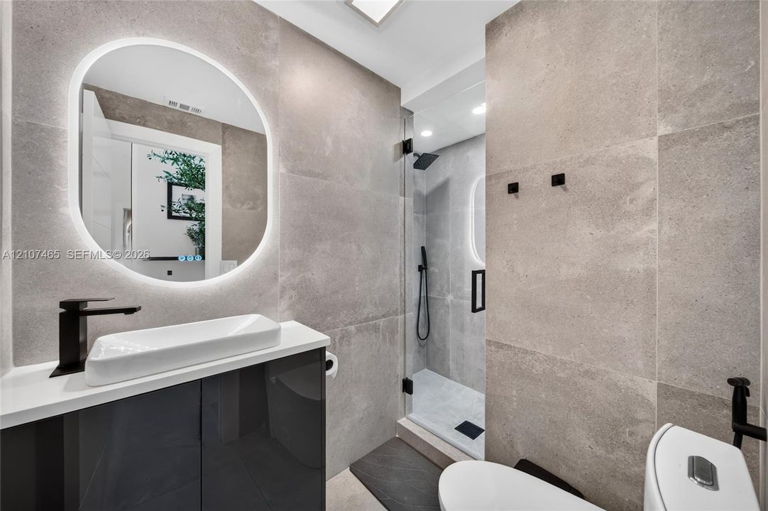 Modern bathroom featuring large-format grey tile wall cladding, a glossy charcoal vanity with a white vessel sink, a backlit oval mirror, and a walk-in shower with a glass enclosure and matte black fixtures