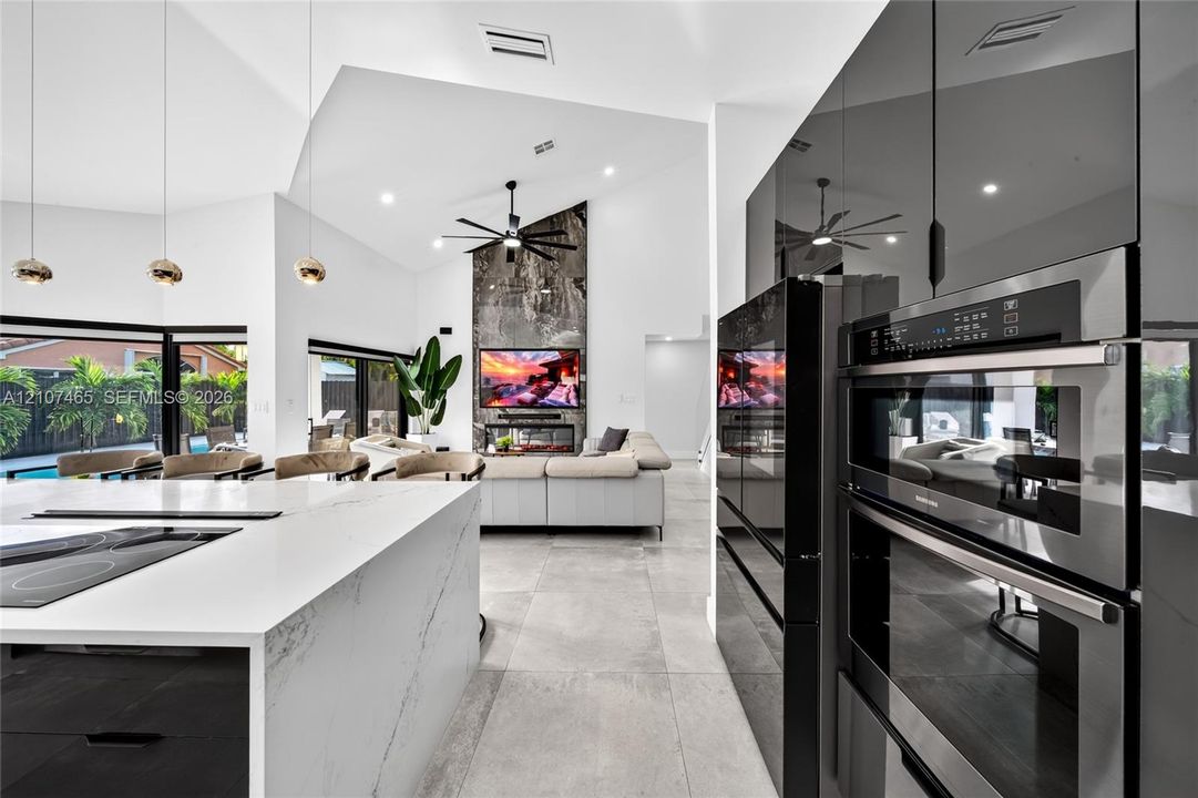 Open-concept kitchen and living area featuring high vaulted ceilings, a central island with waterfall-edge countertop, sleek black built-in wall ovens, large-format tile flooring, and a floor-to-ceiling stone-surround fireplace