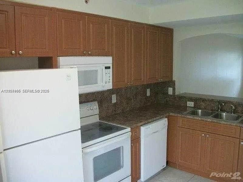 Kitchen featuring medium-tone wood cabinetry, stone-finish countertops, dual-basin stainless steel sink, and white appliances