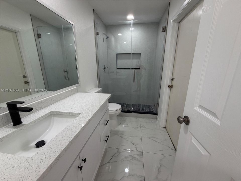 Vanity with quartz countertop, undermount sink, and matte black faucet, featuring large-format tile flooring, a glass-enclosed shower with a built-in recessed niche, and contemporary black cabinet hardware