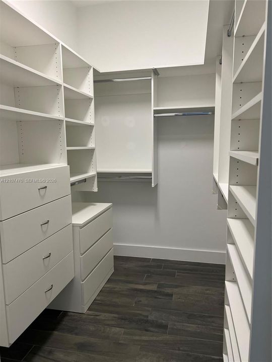 Customized walk-in closet featuring floor-to-ceiling white cabinetry, integrated drawer storage, multiple tiered shelving units, and wood-finish flooring