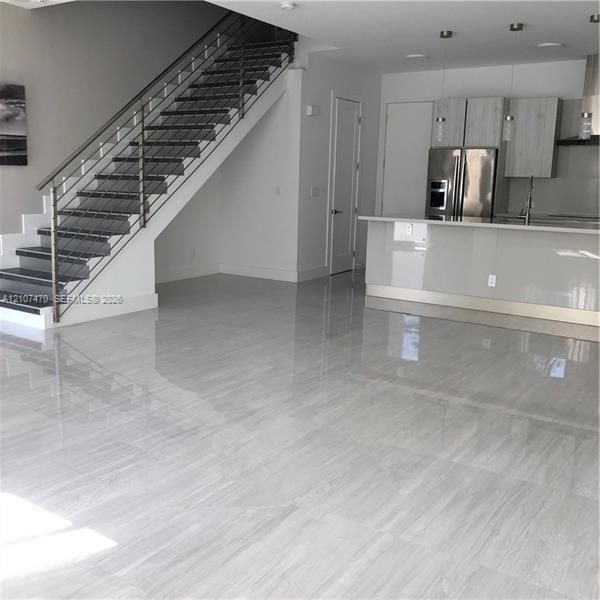 Open-concept interior featuring large-format tile flooring, a modern staircase with a metal cable railing, flat-panel cabinetry, a kitchen island with a stone-finish countertop, and stainless steel appliances