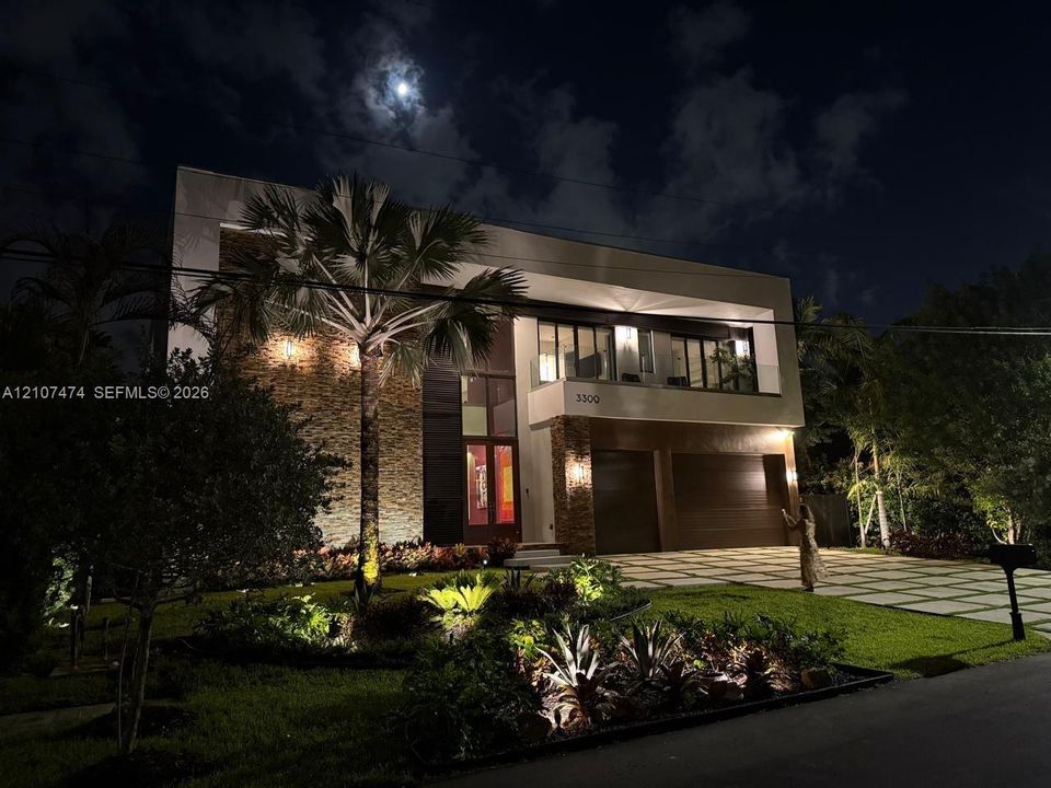 Modern two-story architectural exterior featuring stone facade detailing, integrated landscape lighting, glass-railed upper balcony, and a multi-car garage with wood-finish doors