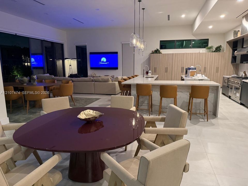 Open-concept interior featuring large-format tile flooring, recessed ceiling lighting, a waterfall-edge kitchen island, full-height light wood-finish cabinetry, and glass pendant light fixtures