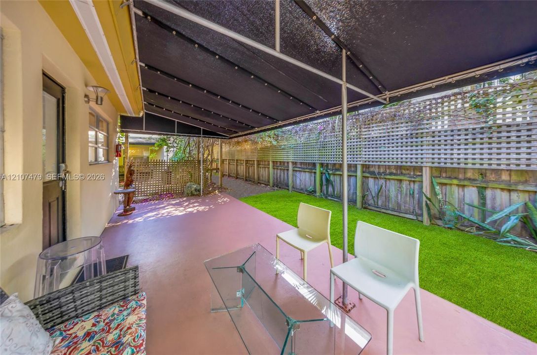 Cottage guest house, Covered patio featuring an extended black fabric awning, solid surface flooring, artificial turf lawn, and perimeter wood privacy fencing