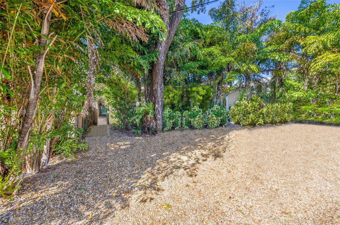 Gate entrance to Cottage guest house, Ground-level exterior area featuring gravel surfacing, mature trees, dense perimeter foliage, and a paved walkway