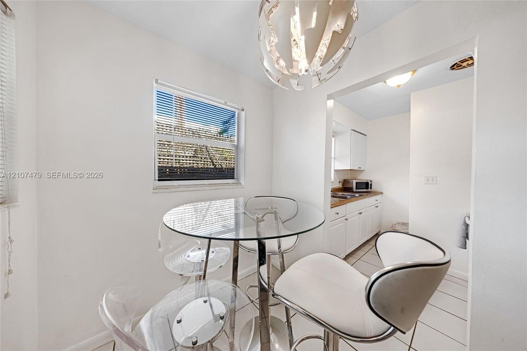 Cottage / guest house, Dining area featuring a modern sculptural pendant light fixture, ceramic tile flooring, and a window with blinds, opening into a kitchen equipped with white cabinetry and stone countertops