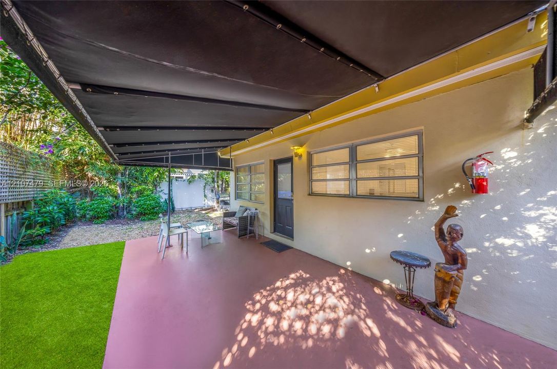 Cottage guest house Entrance, Covered concrete patio featuring a retractable fabric awning, horizontal sliding windows, and a fenced backyard with artificial turf