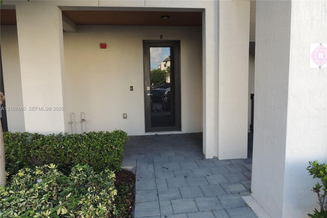 Covered entryway featuring a dark-framed glass door, exterior stucco finish, paver-stone walkway, and manicured shrubbery