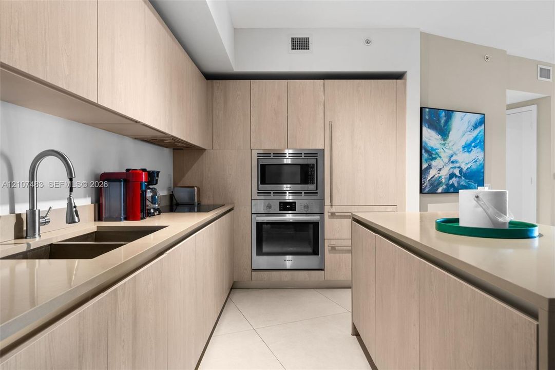 Kitchen featuring light-finish wood cabinetry, integrated stainless steel oven and microwave, stone-finish countertops, and an undermount dual-basin sink with a high-arch faucet