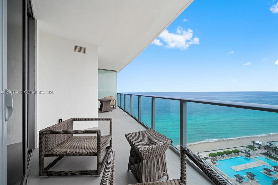 Covered balcony featuring a sleek glass-panel railing, neutral tiled flooring, and expansive ocean and coastal scenery