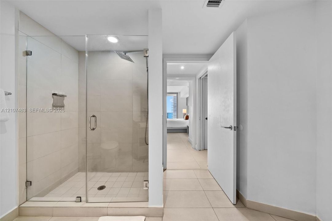 Walk-in shower featuring a glass enclosure, large-format tile surround, rainfall shower head, and built-in metal shelving, complemented by light-toned tile flooring