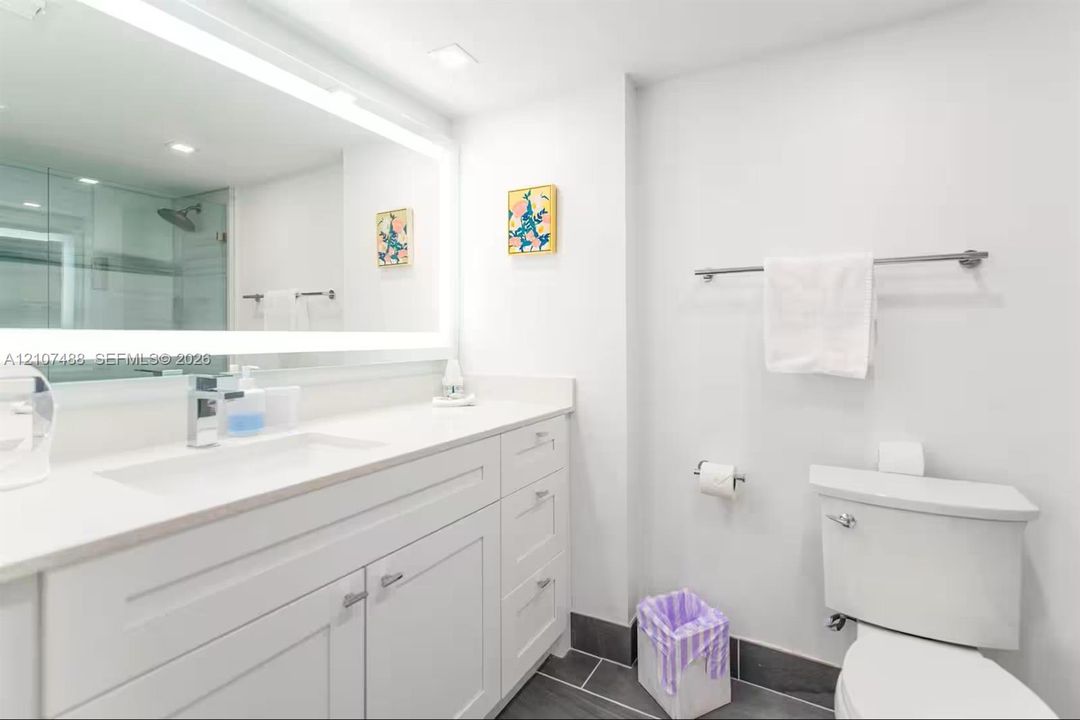Bathroom featuring a vanity with white cabinetry and a solid-surface countertop, an integrated rectangular sink, a large wall-mounted LED-backlit mirror, and dark tile flooring