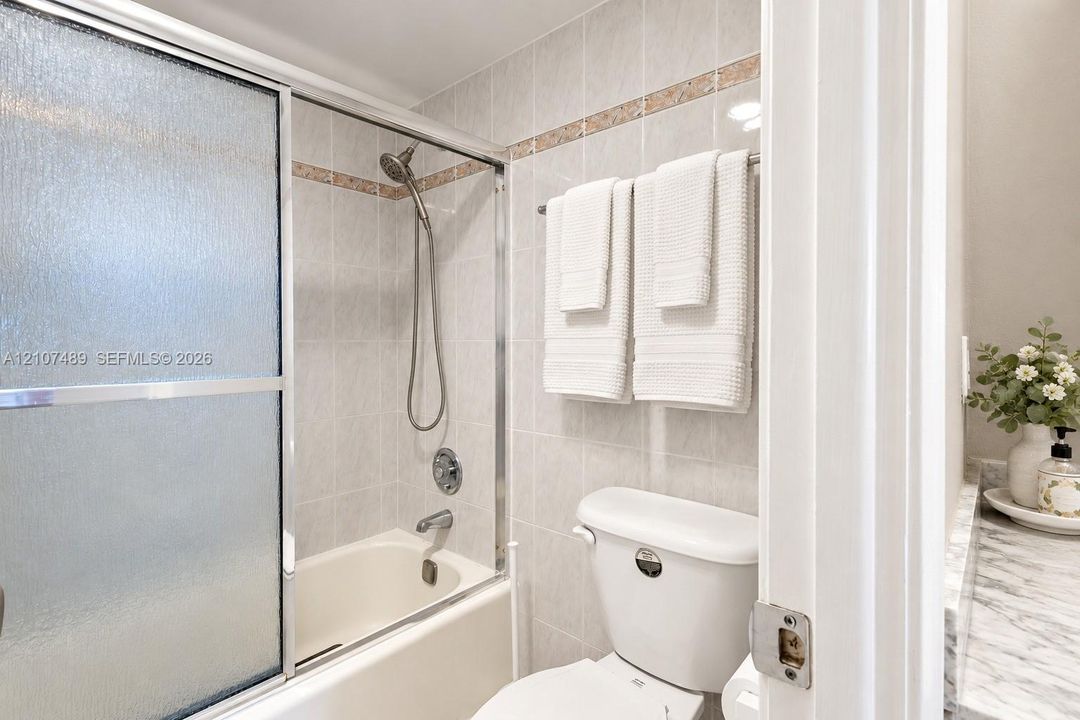 Bathtub with glass sliding doors, integrated handheld shower fixture, light-toned wall tile with decorative accent border, and stone-finish vanity countertop