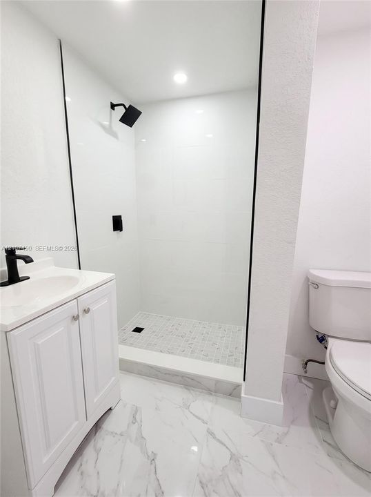 Walk-in shower with floor-to-ceiling white tile, glass enclosure, and matte black hardware, accompanied by a white vanity with integrated sink and stone-patterned flooring