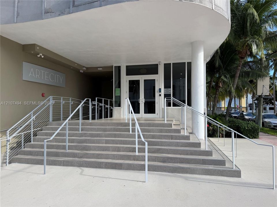 Building entrance featuring a concrete staircase with metal railings, a covered porch, glass-paned double entry doors, and a concrete-paved approach