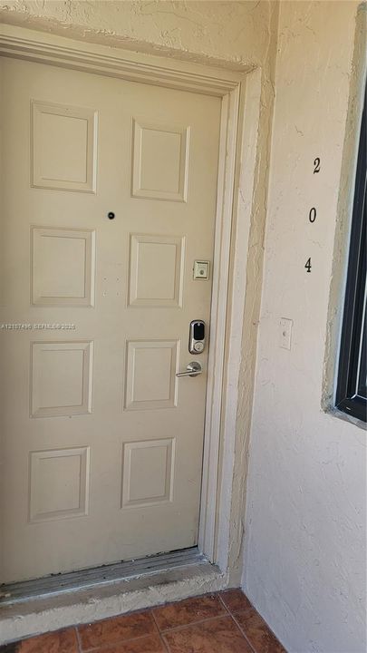 Eight-panel entry door featuring a digital smart lock, integrated peephole, and metallic lever handle, set against textured stucco exterior walls and tile flooring