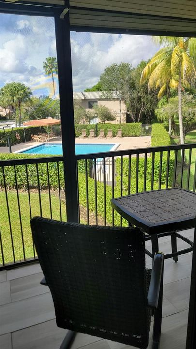 Screened balcony featuring metal railings and tile flooring, overlooking a community swimming pool, concrete deck, and perimeter hedges