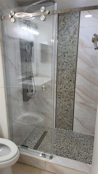 Walk-in shower featuring a sliding glass door, multi-jet shower panel, stone-tiled floor, and a decorative vertical stone mosaic inlay