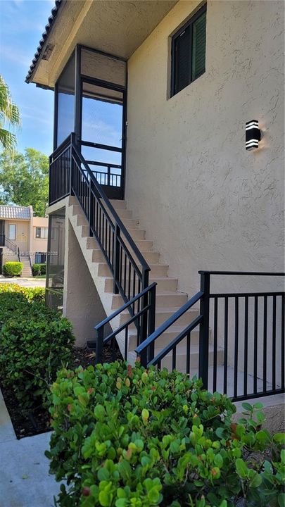 Exterior staircase with black metal railings, screened-in entry landing, stucco siding, and wall-mounted exterior lighting